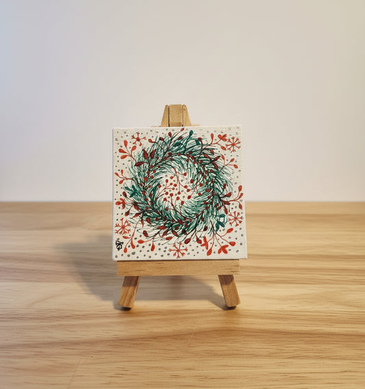Small painting of a green wreath with red flowers on a white background, displayed on a wooden easel.