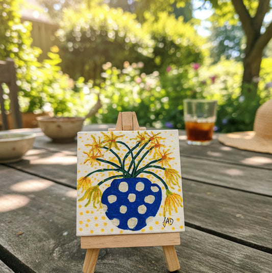 Small painting of a blue vase with yellow flowers on a white background, displayed on a wooden easel.