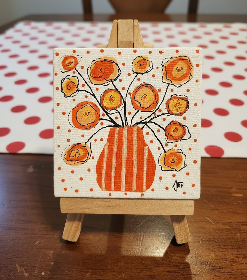Small painting of a vase with orange flowers on a white background with orange dots, displayed on a wooden easel.